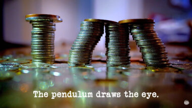 Image of four stacks of coins, arranged to resemble the letters 'TM' (a reference to the 'Put the largest numeric value of money in the floating bowl' task), with the episode title, 'The pendulum draws the eye', superimposed on it.