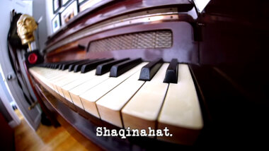 Distorted wide angle image of a piano keyboard, with a Taskmaster trophy visible at its far end (a reference to the 'Write and perform lyrics for the Taskmaster theme tune' task, and possibly also the 'Supply a soundtrack for a film' task), with the episode title, 'Shaqinahat', superimposed on it.
