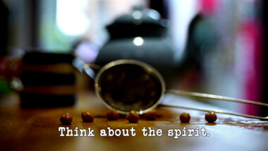 Image of six chickpeas on a wooden surface, in front of a small sieve (a combined reference to both the 'Do the most preposterous thing with a chickpea' task and the 'Make a cup of tea' task), with the episode title, 'Think about the spirit', superimposed on it.