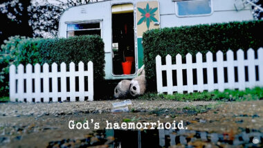 Image of the feet of a giant teddy bear, poking out from behind the low hedge around the Taskmaster's caravan (a reference to the 'Fell the coconut, then move the drinks' task), with the episode title, 'God’s haemorrhoid', superimposed on it.