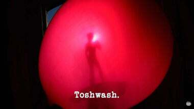 Image of the shadow of a figurine against the surface of an inflated red balloon (a combined reference to the 'Drop a water balloon as far as possible without it bursting' task, and the 'Persuade the security guard to look in your bag' task), with the episode title, 'Toshwash', superimposed on it.