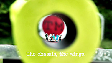 Image of miniature plastic figures of the series' contestants, viewed through the centre of a roll of yellow toilet paper (a reference to the 'Land loo roll in the toilet' task), with the episode title, 'The chassis, the wings', superimposed on it.