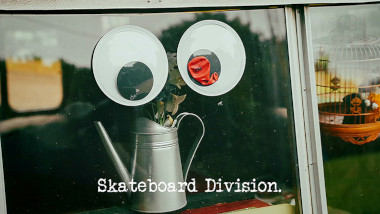 Photo of two giant googly eyes stuck to the window of the Taskmaster caravan, one of which has a deflated red Taskmaster balloon attached to it (a reference to both the 'Make a cool but scary googly-eyed gang' and 'Burst the balloon without looking at it' tasks), with the episode title, 'Skateboard Division', superimposed on it.