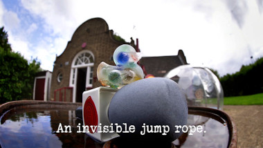 Photo of ice containing coloured balls, frozen in the shape of a rubber duck, perched on top of a microphone on an oil drum in the driveway of the Taskmaster house (referencing the 'Get the most liquid in this can' and 'Achieve a really tricky thing after commentating on the achievement' tasks), with the episode title, 'An invisible jump rope', superimposed on it.