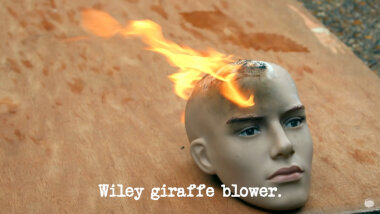 Image of a mannequin's head, set aflame, on top of a wooden board on the ground (taken during Bob Mortimer's attempt at the 'Perform a miracle' task), with the episode title, 'Wiley giraffe blower', superimposed on it.