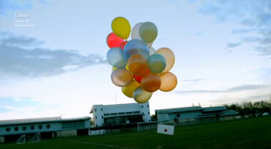 Image of the task brief suspended below a bunch of balloons in the middle of a football pitch.
