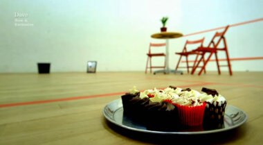 Image of a squash court, with a tray of cupcakes in the foreground, and a waste paper bin, framed picture, and table (with potted plant) and chairs in the background.