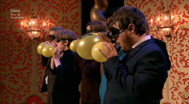 Image showing the contestants lined up on stage, in the process of inflating their balloons, while wearing blindfolds.