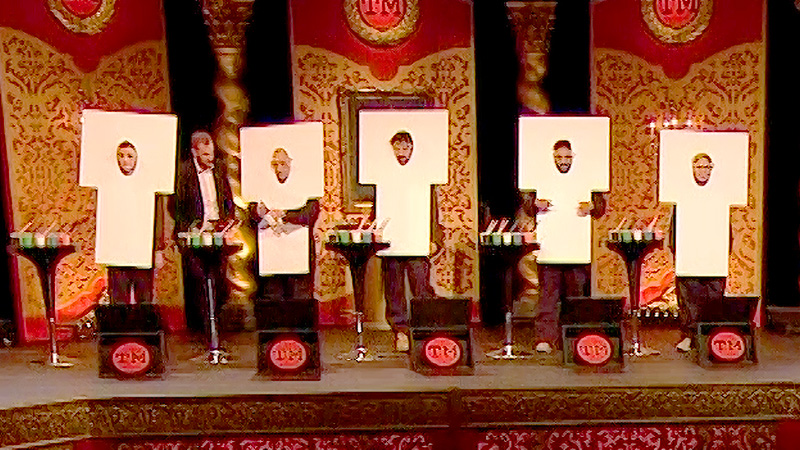 The cast of Taskmaster UK series 5 on stage with their heads poking through canvases on which they have painted. Some contestants have painted their faces as well. Three contestants are painted to look like carrots.