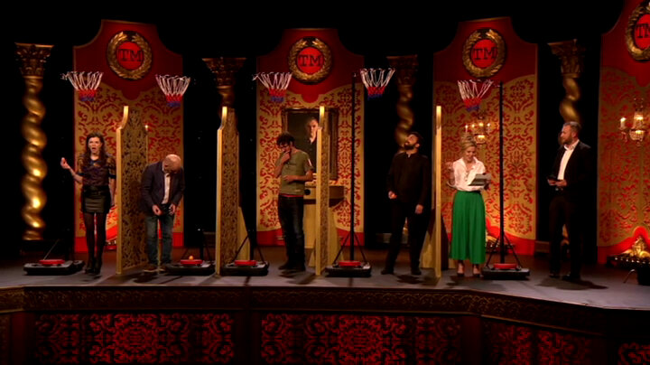 The cast of UK series 5 stands on stage with dividers between them and a basketball hoop over each of their heads. Alex Horne looks on.