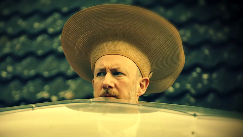 Alex Horne in a large-brimmed straw hat, peering over the geodesic dome.