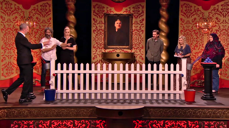Alex Horne walks in front of white picket fence with a bucket on each end. The teams confer at opposing plinths.