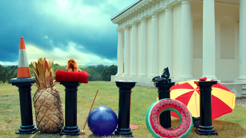 Five plinths with various objects set on and around them, including: a traffic cone, a croquet mallet, a pair of rollerskates, and watermelon pool float.